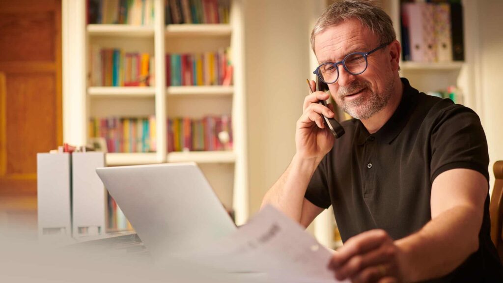 man sitting at a table on the phone, looking at a sheet of paper in his hands